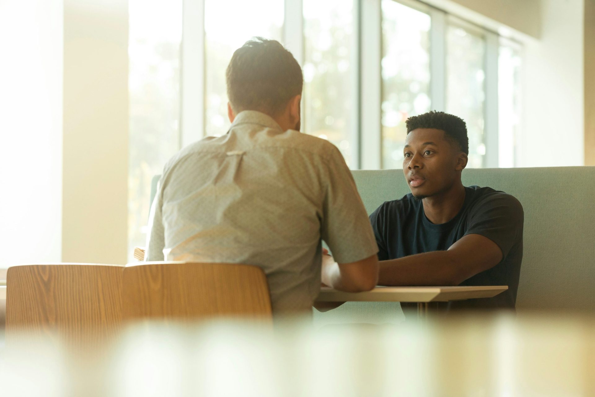 two men sitting at a table by a window having a discussion